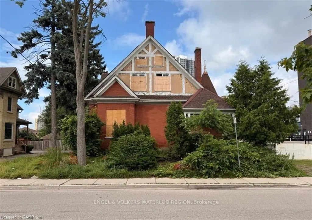 Exterior view of a red brick house with boarded-up windows and a gable roof, surrounded by trees and greenery.