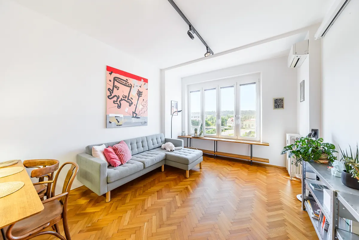 Bright living room with herringbone wood floors, a gray sectional sofa with pink pillows, and a large abstract painting on a white wall.