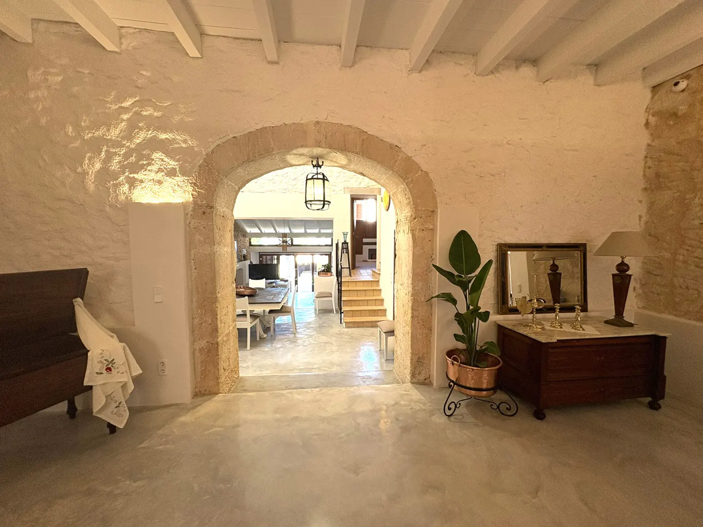 Interior view of a room with stone walls and a stone archway leading to a dining area and stairs. A wooden chest and plant are visible.