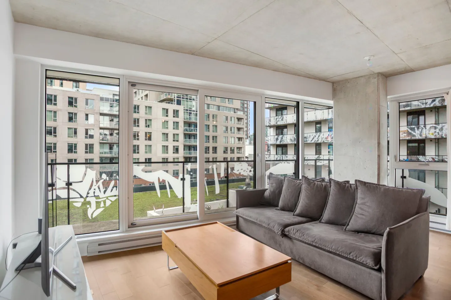 Modern living room with a grey sofa, wooden coffee table, and floor-to-ceiling windows overlooking city buildings.
