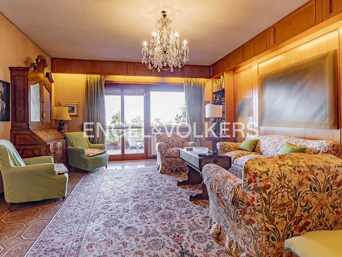 Classic living room with floral sofas, green armchairs, a crystal chandelier, and wood-paneled walls overlooking a balcony.