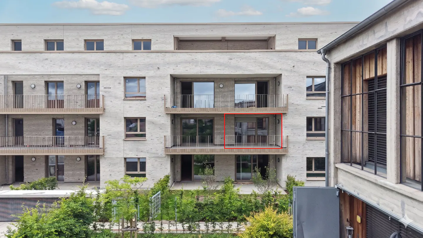Exterior view of a modern apartment building with balconies, light brick facade, and green landscaping. One balcony is highlighted in red.