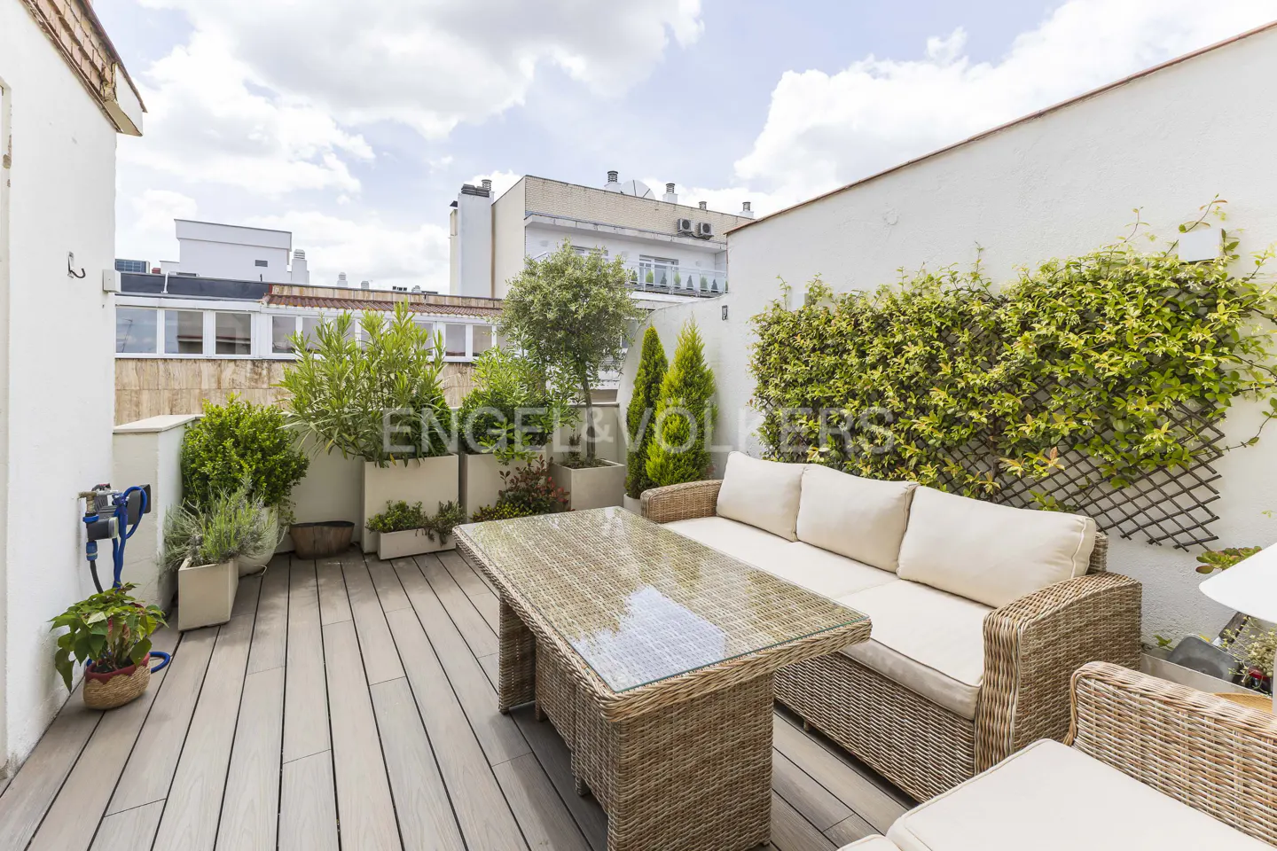 Outdoor patio with wicker furniture, including a sofa and table, on a gray wood deck. Green plants and vines adorn the white walls.