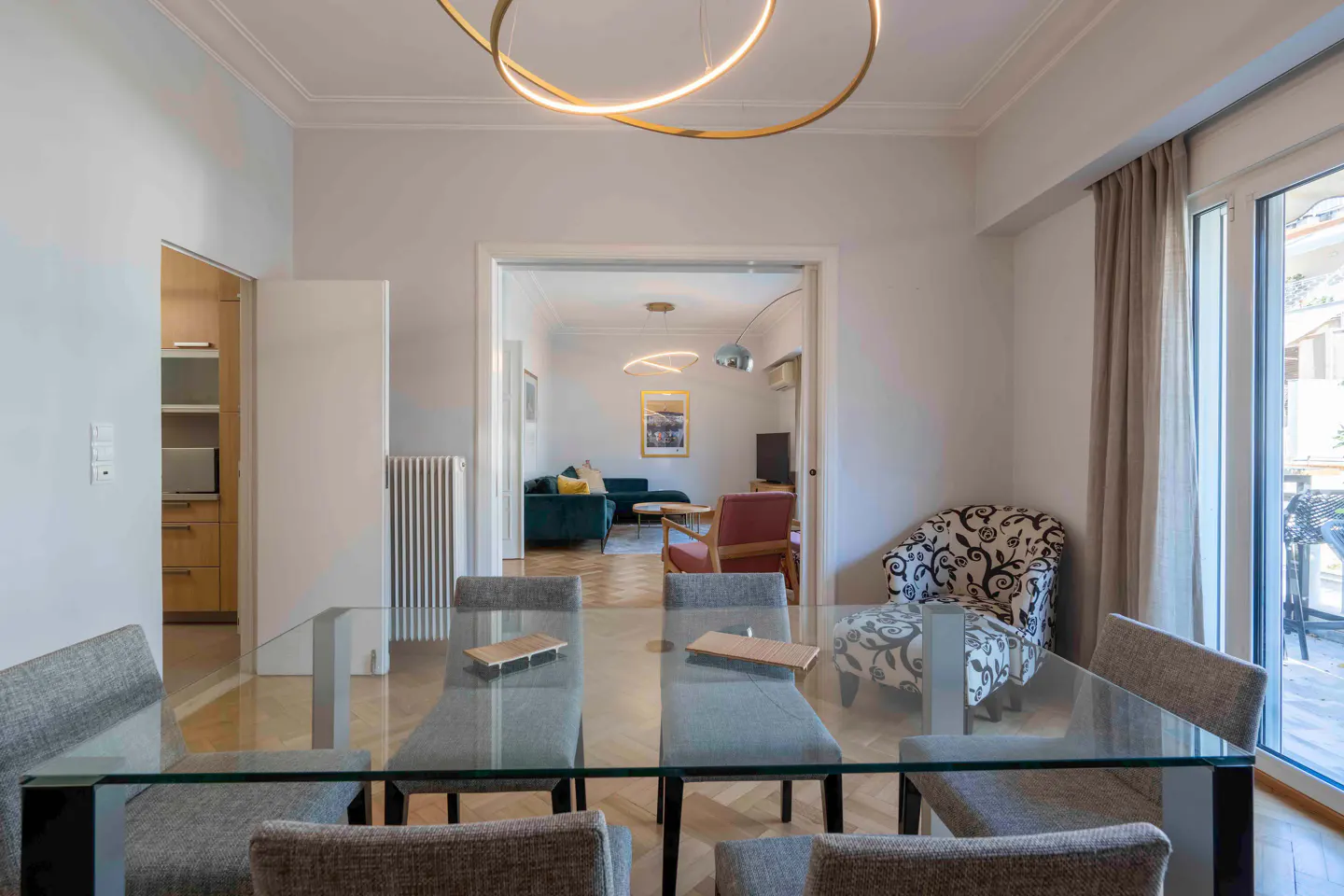 A bright, modern dining room with a glass table and gray chairs, leading to a living room with a teal sofa.