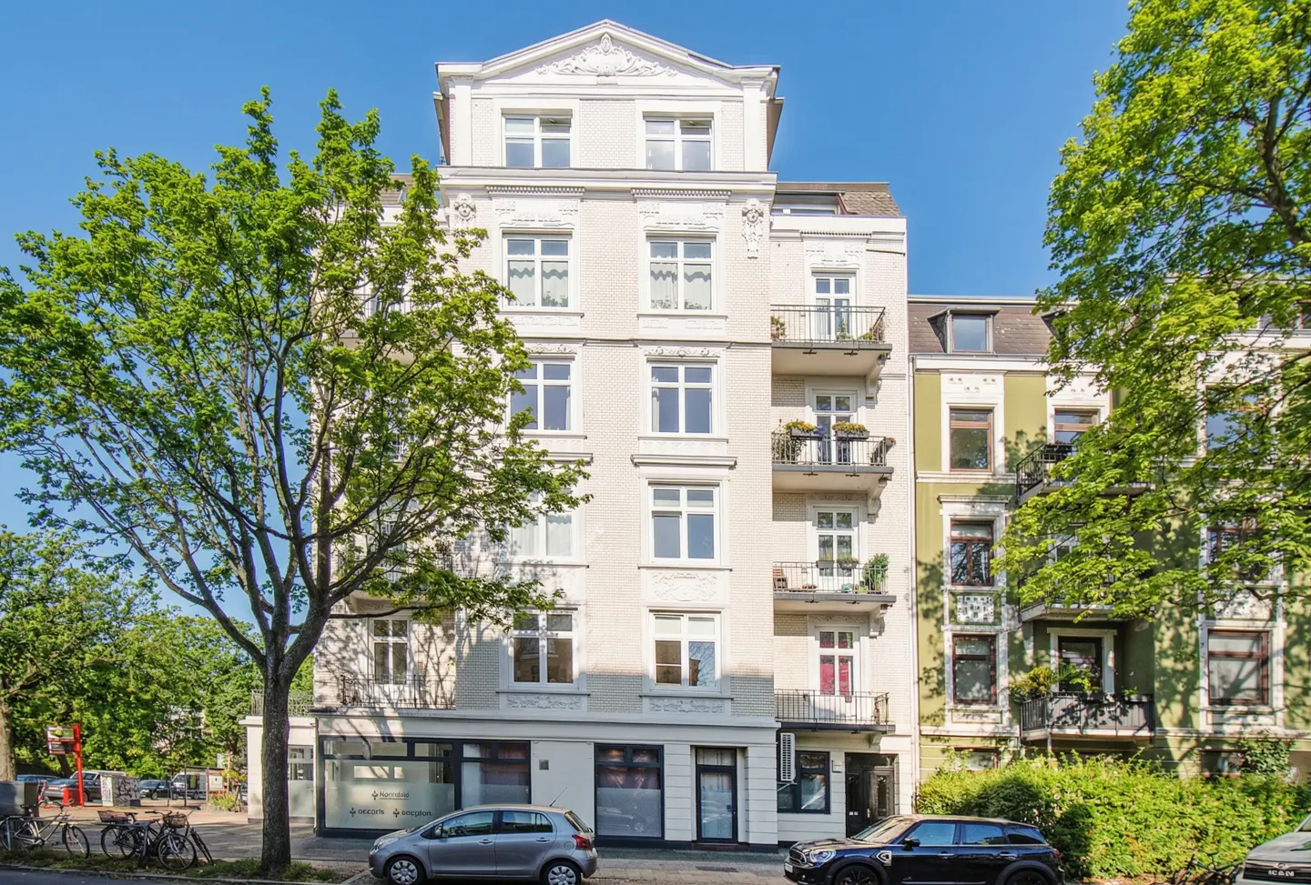 Exterior view of a five-story white brick apartment building with balconies, cars parked on the street, and green trees.