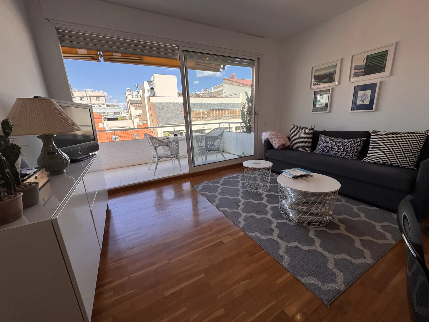 Living room with wood floors, a gray sofa with patterned pillows, and a gray rug. A balcony with outdoor furniture is visible through sliding glass doors.