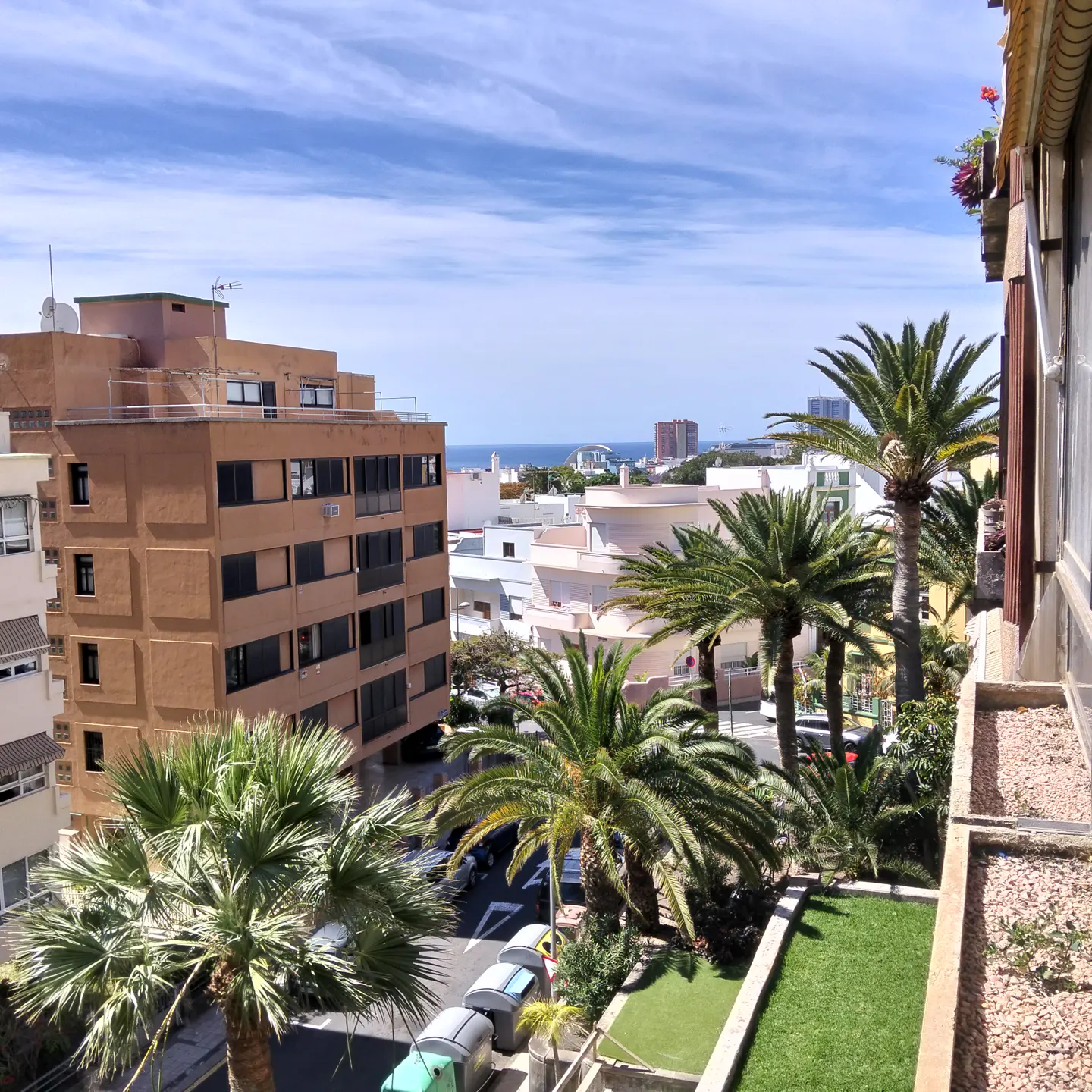 View from a balcony overlooking a city street with palm trees, buildings, and the ocean in the distance under a blue sky.