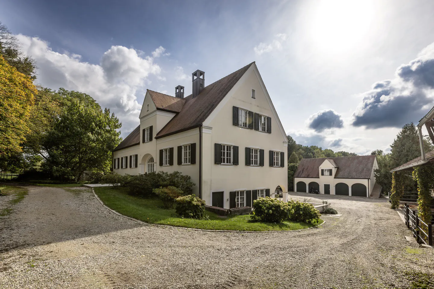 Exterior view of a large, two-story cream-colored house with black shutters and a brown roof, surrounded by trees and a gravel driveway.