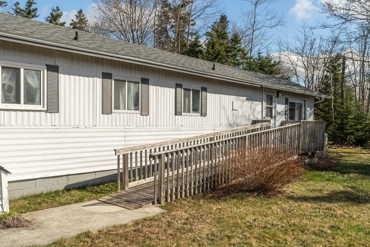 Exterior view of a white, one-story house with a wooden ramp leading to the entrance. Gray shutters adorn the windows. Trees in the background.