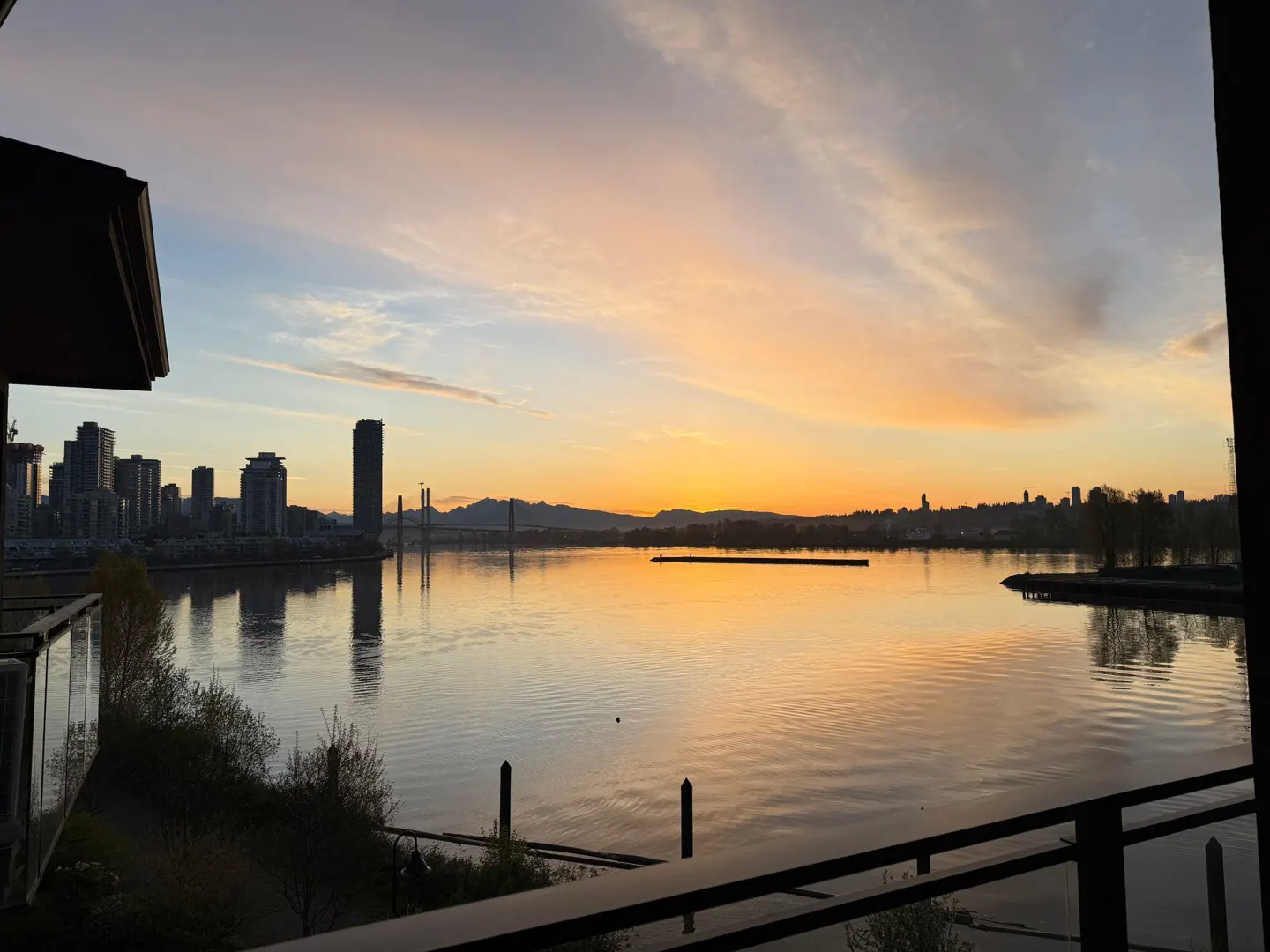 Sunset view from a balcony overlooking a calm body of water, with a city skyline in the distance. The sky is a mix of orange, pink, and blue.