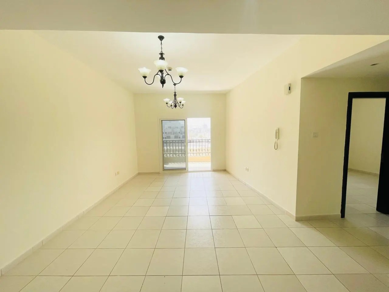 Empty apartment with beige walls, tiled floor, and balcony. Two chandeliers hang from the ceiling. A doorway leads to another room.