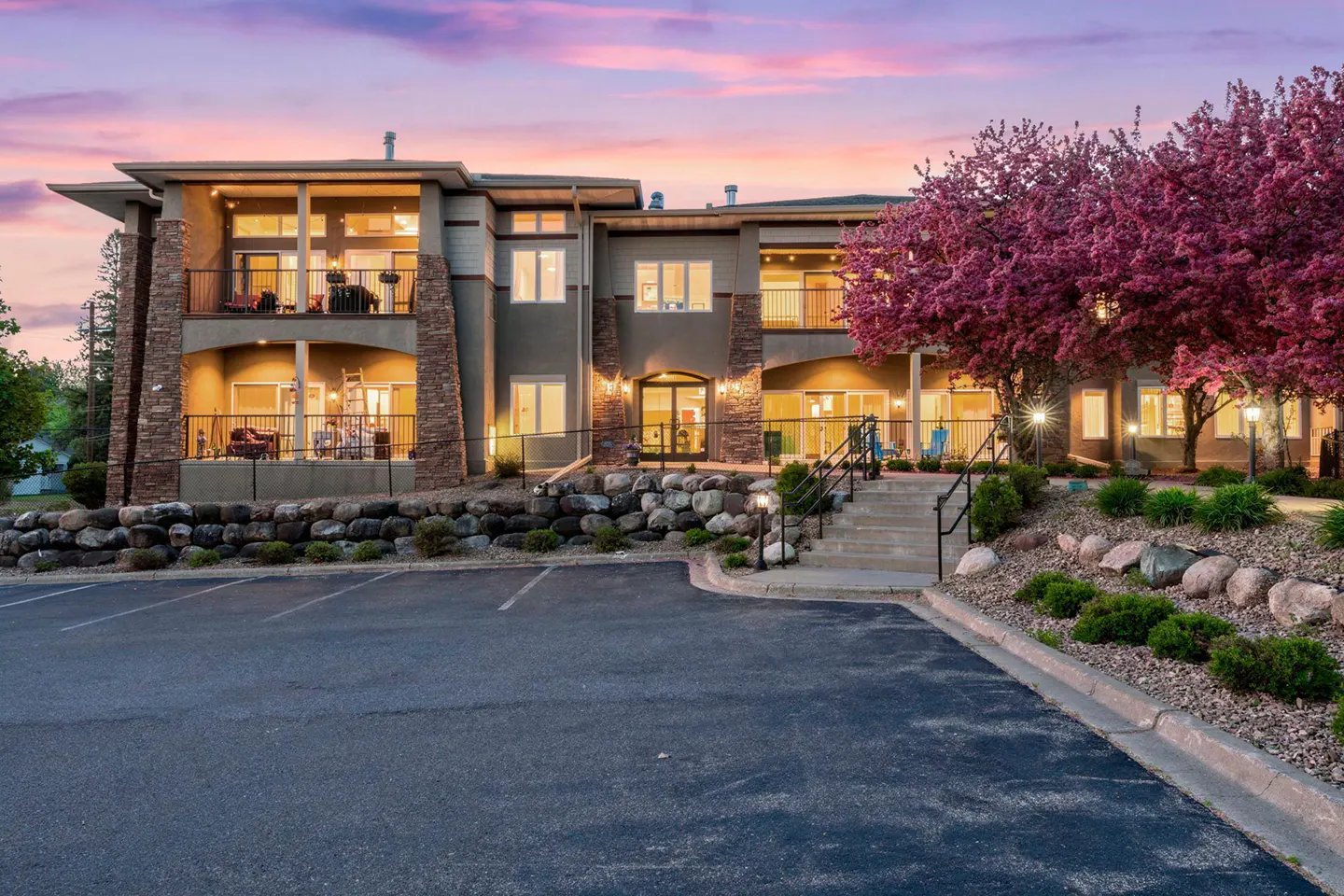Two-story apartment building with balconies, stone accents, and a pink sunset sky. A parking lot is in the foreground.