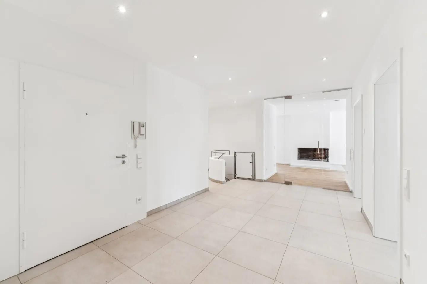 Bright, empty room with white walls, beige tile floor, and recessed lighting. A glass door leads to a room with a fireplace.