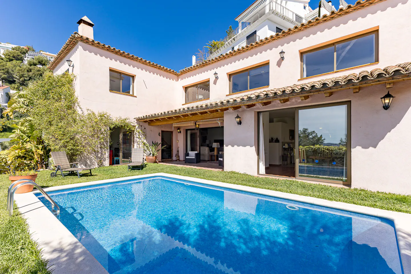 Exterior view of a pink house with a blue swimming pool and green grass. The house has brown window frames and a brown roof.