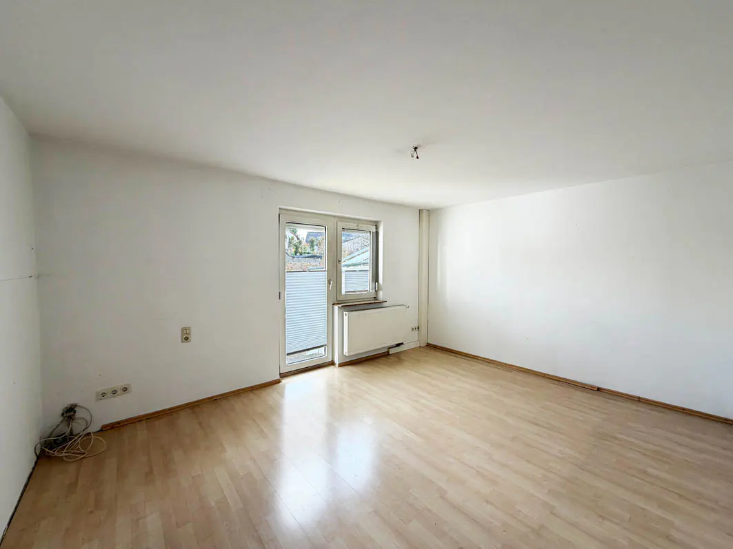 Empty room with light wood floor, white walls, and a door to a balcony with blinds. A radiator is under the door.