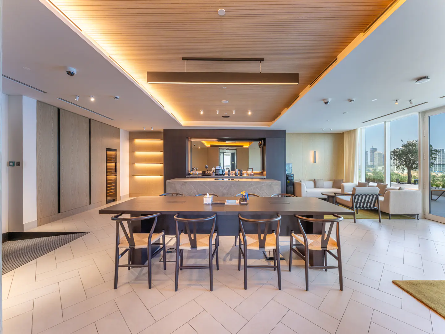 A modern lounge area with a long dark table and chairs, a kitchenette, and seating near large windows with a city view.