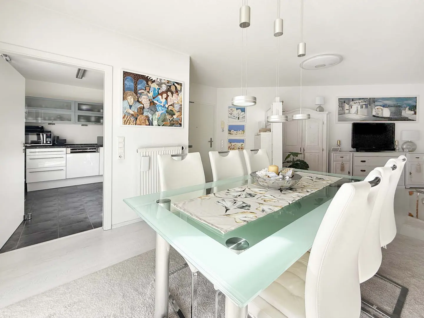 Bright dining room with a glass table, white leather chairs, and modern pendant lights. Kitchen visible through doorway. Artwork on walls.