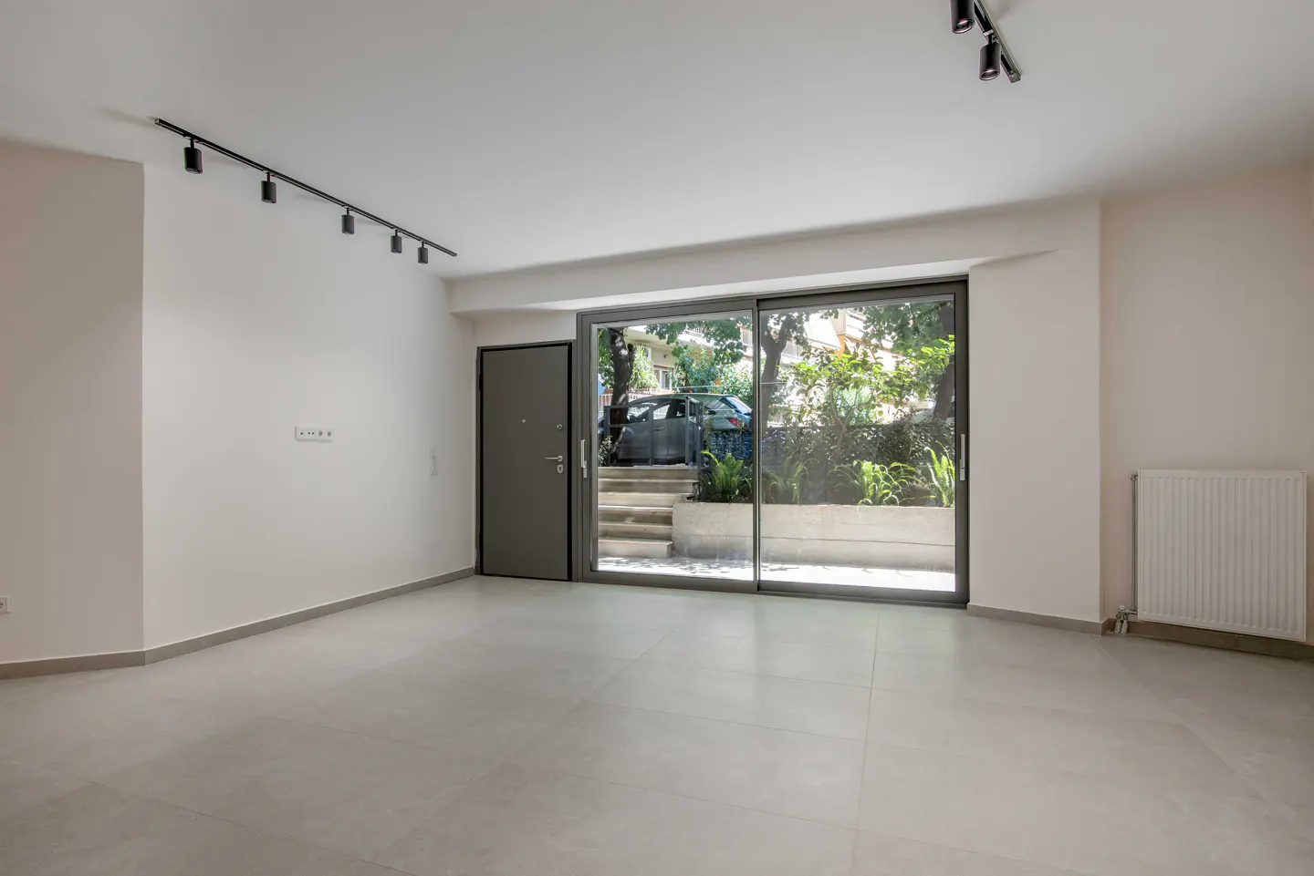 Empty room with white walls, light tile floors, track lighting, and a sliding glass door leading to a patio and garden.