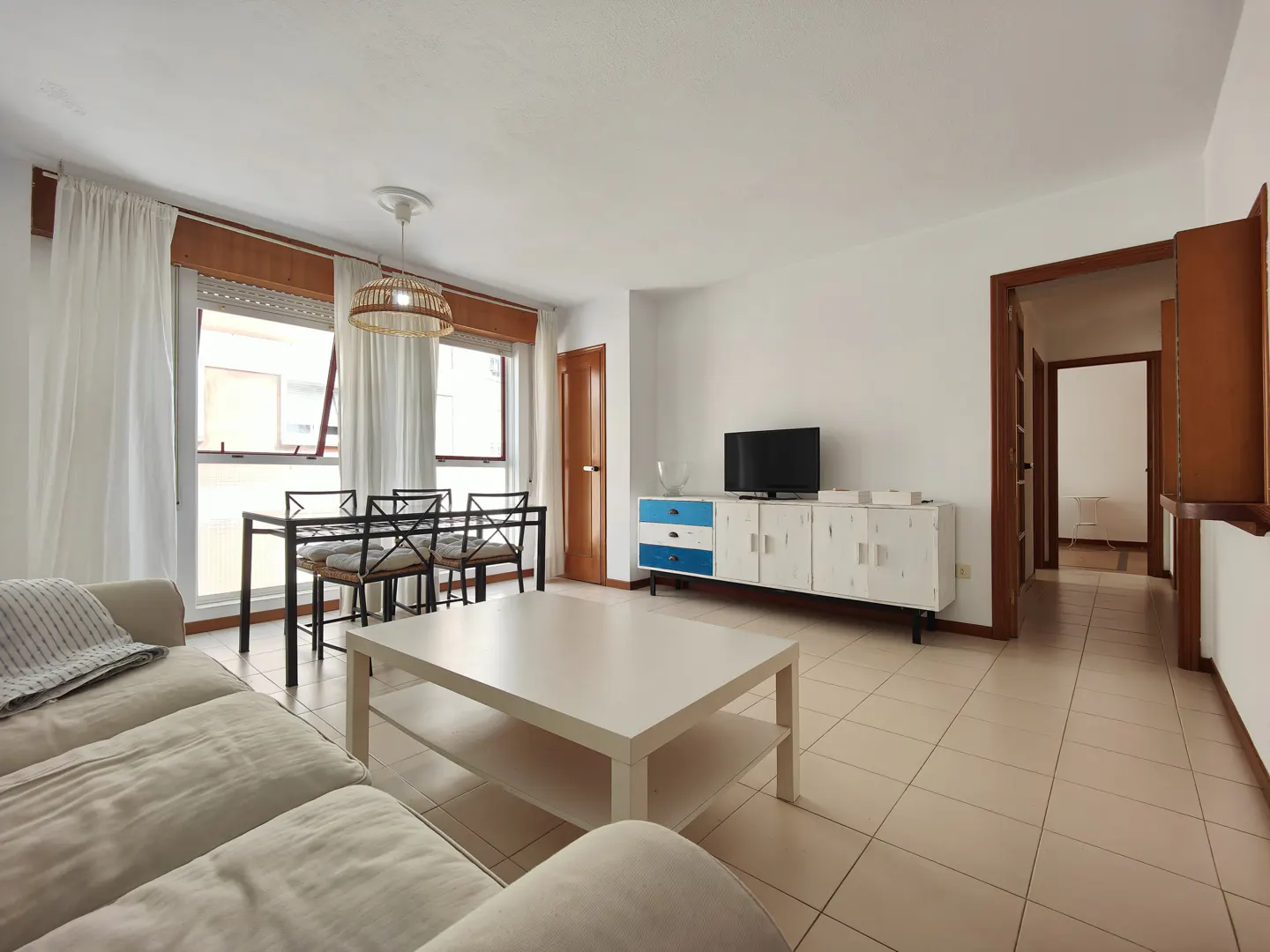 Bright living room with beige sofa, white coffee table, dining table, and white cabinet with blue drawers. Tiled floor.
