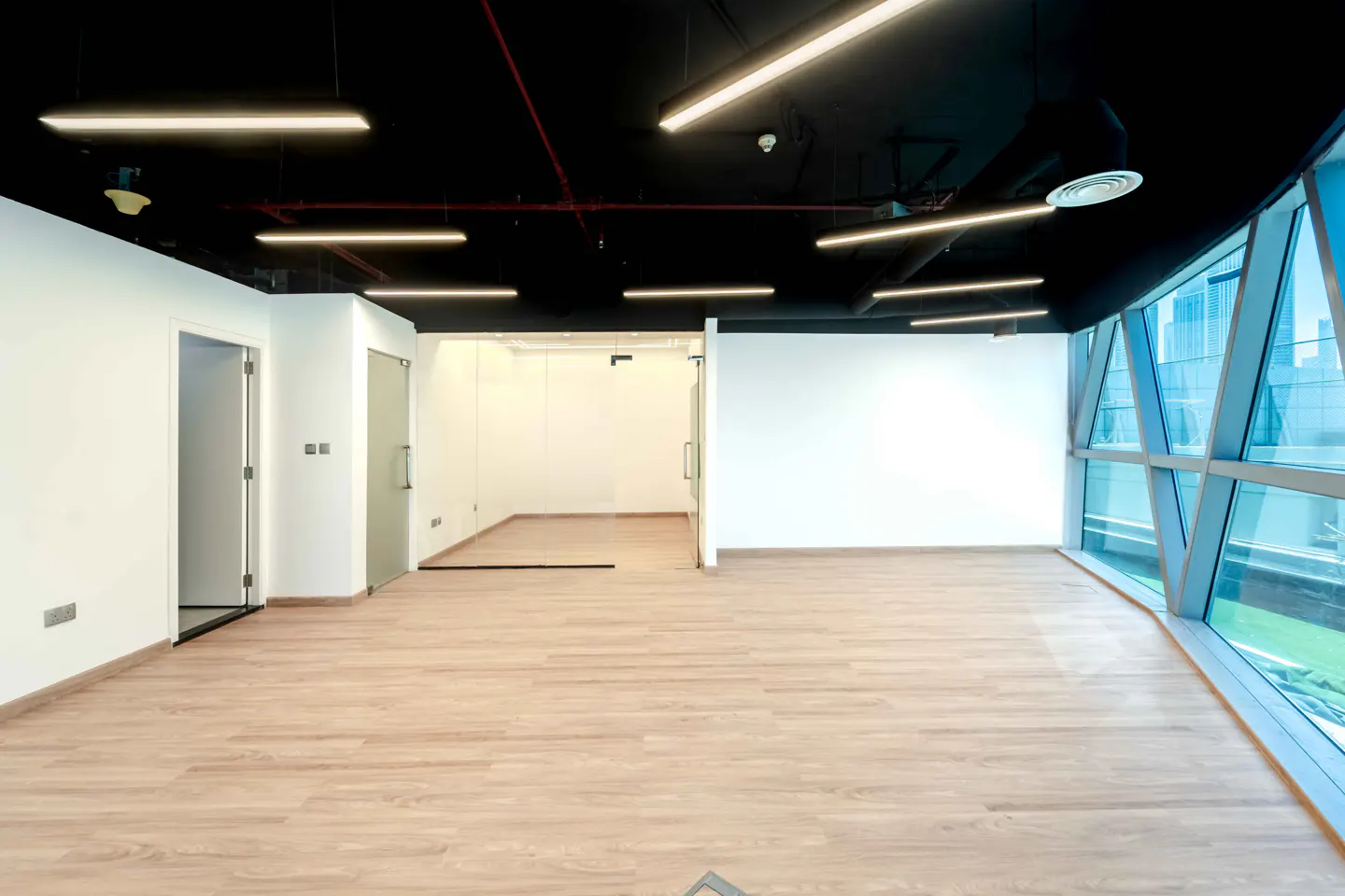 Empty office space with light wood floors, white walls, and a black ceiling with linear lights. Large windows offer a city view.