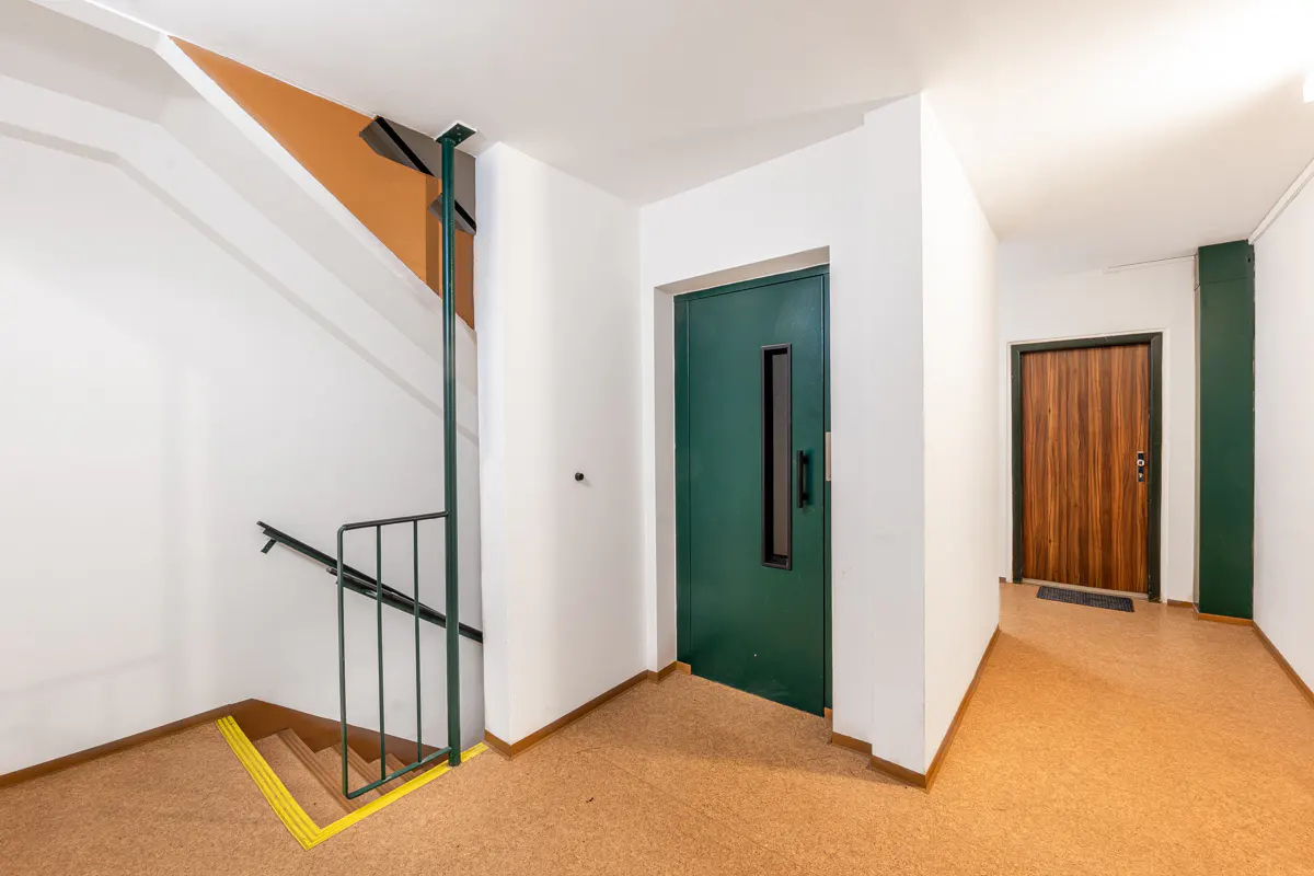 Hallway with stairs, a green elevator door, and a wood door. The walls are white, and the floor is brown.