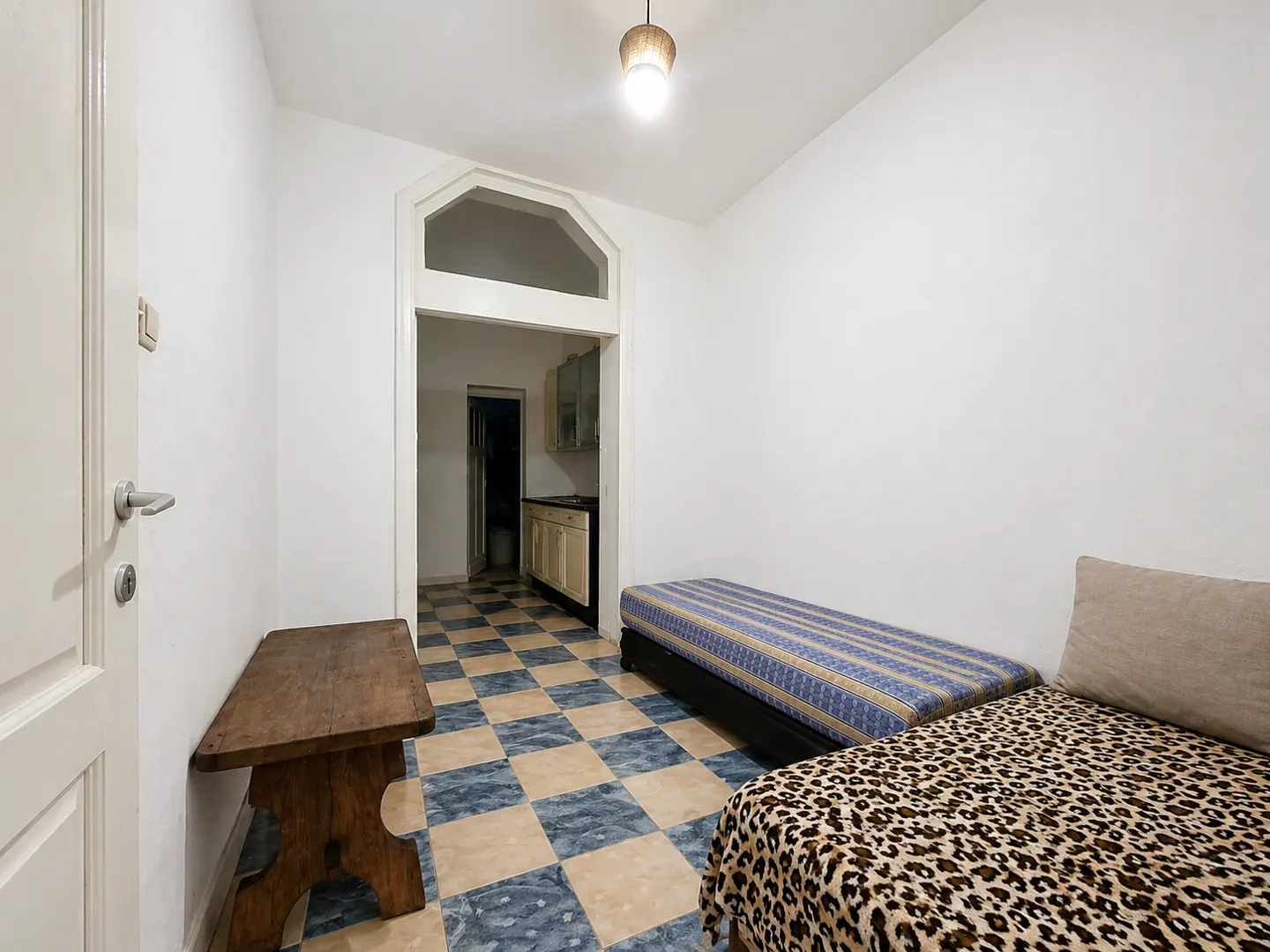 A small room with two beds, one with a blue striped cover and the other with a leopard print, and a wooden bench. A kitchen is visible through an arched doorway.