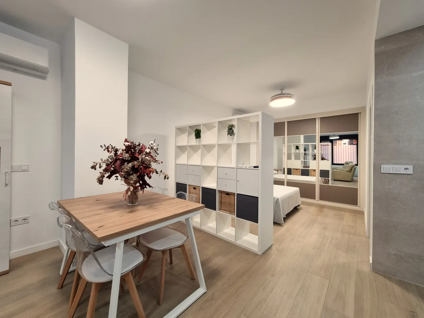 Bright studio apartment with wood floors. A dining table with flowers sits near a white shelving unit dividing the living space from the bedroom.
