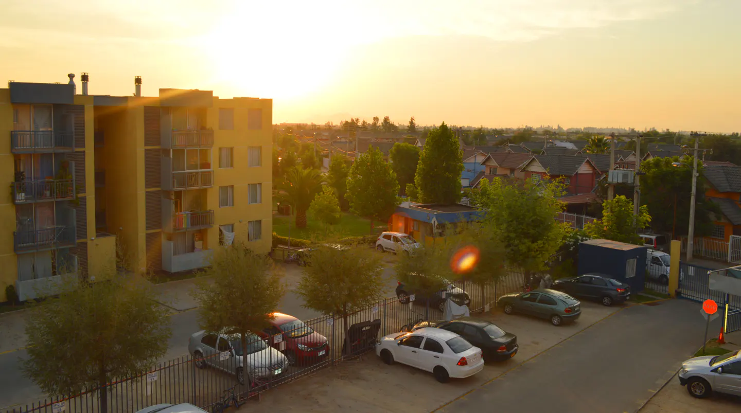 High-angle view of a yellow apartment building and parking lot at sunset, with cars parked among trees and houses nearby.