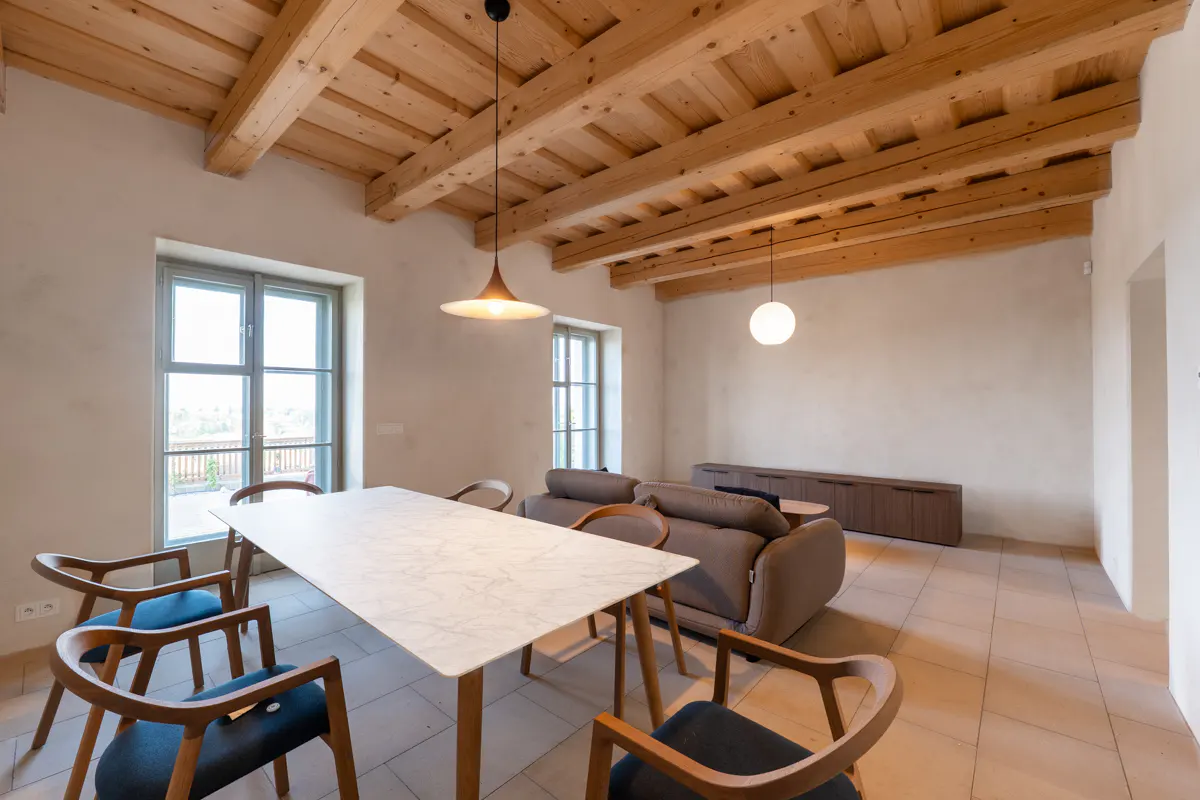 Bright, open-plan living and dining area with wooden beamed ceiling, marble table, and modern furniture. Natural light streams through the windows.