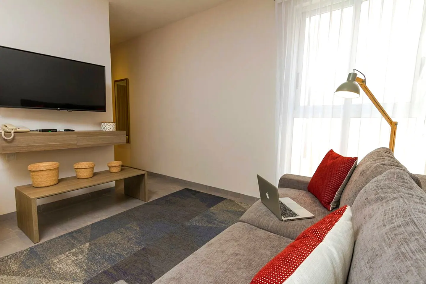Living room with a gray sofa, red pillows, and a laptop. A TV is mounted on the wall above a wooden shelf with baskets. A patterned rug covers the floor.