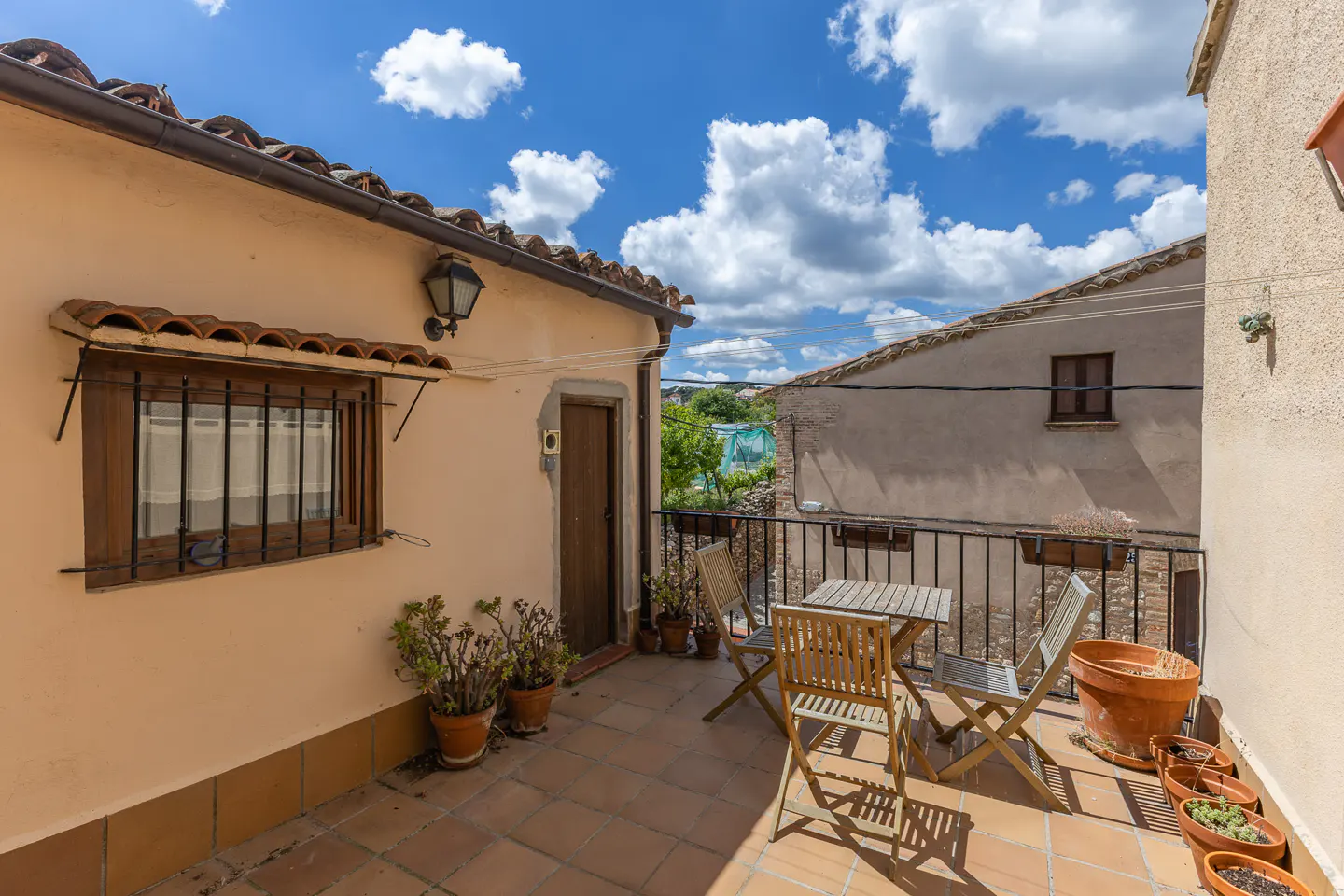 Outdoor patio with terracotta tile, wooden table and chairs, potted plants, and a view of a blue sky with white clouds.