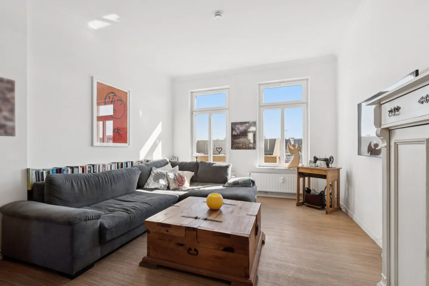 Bright living room with a grey sectional sofa, wooden trunk coffee table, and two windows overlooking a sunny exterior.