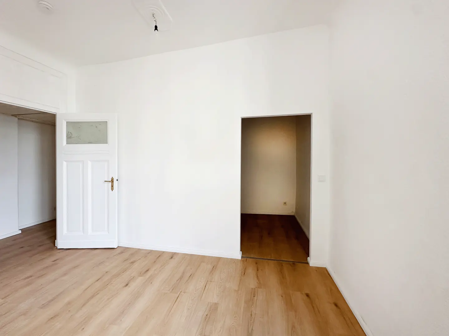 Empty room with white walls, light wood flooring, a white door with a glass pane, and an open doorway to another room.