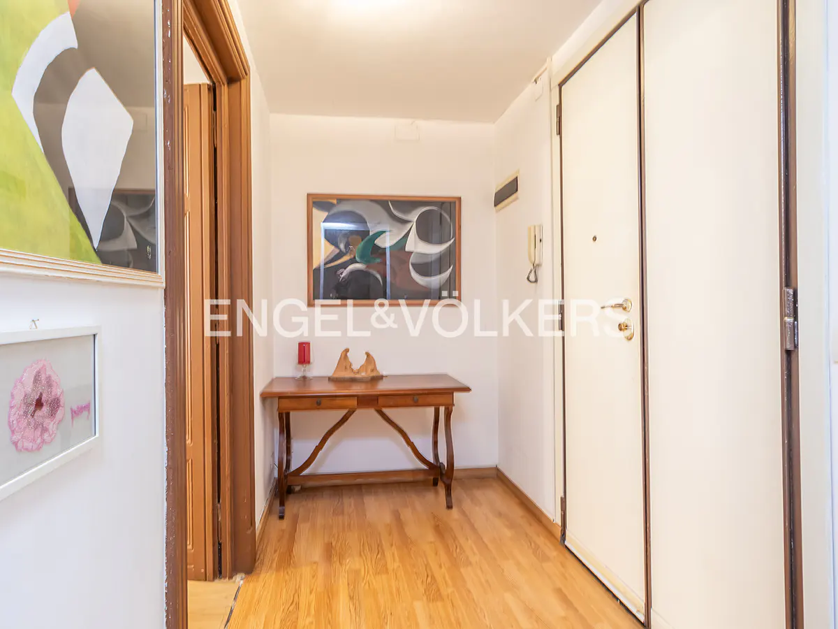 Apartment entryway with wood floors, white walls, and a wooden table with art above it. A white door is on the right.
