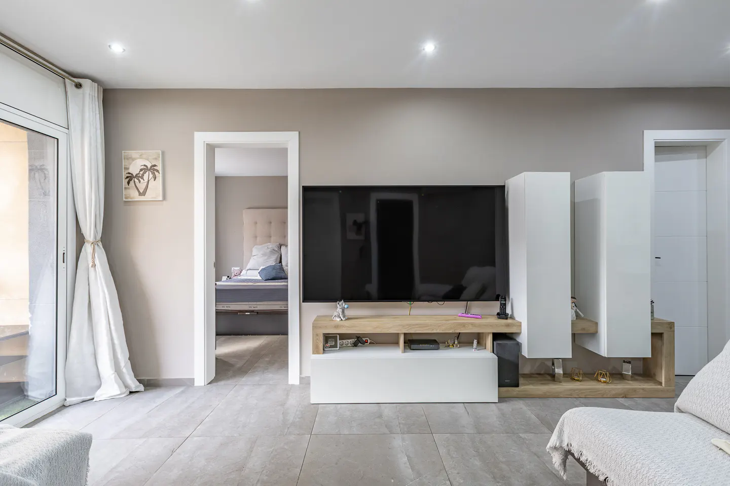 Modern living room with a wall-mounted TV, light wood and white media console, and a view into a bedroom with a bed.