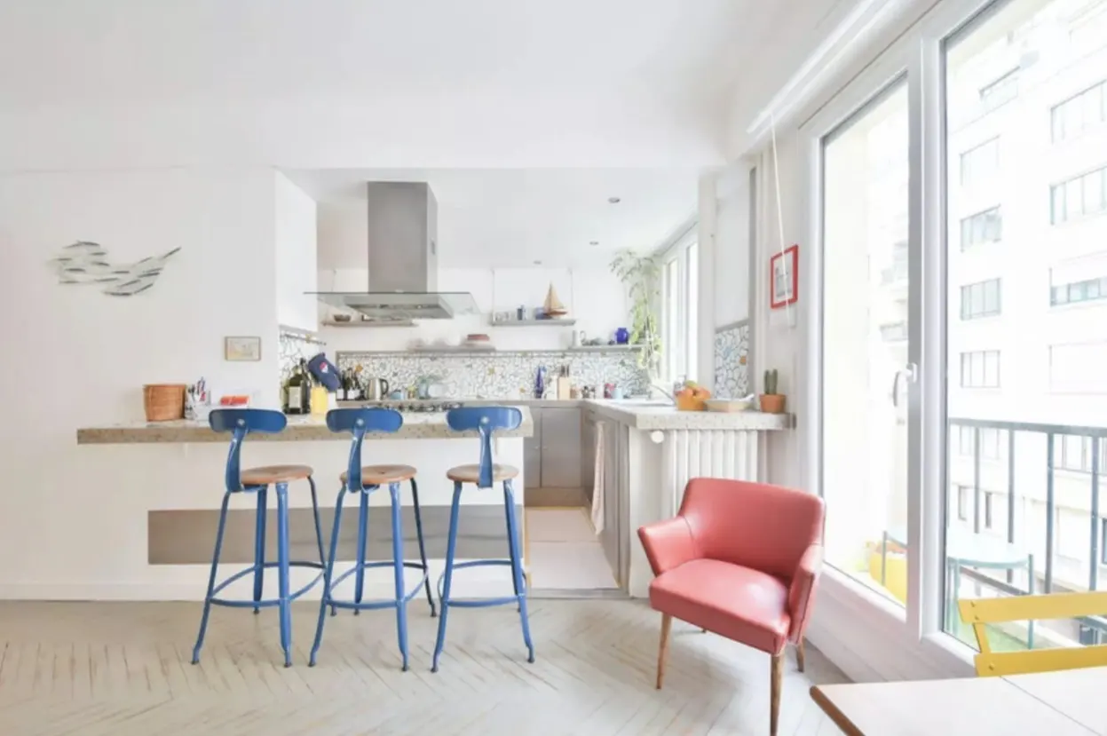 Bright, modern kitchen with blue stools at a counter, a pink chair, and a balcony with a city view.