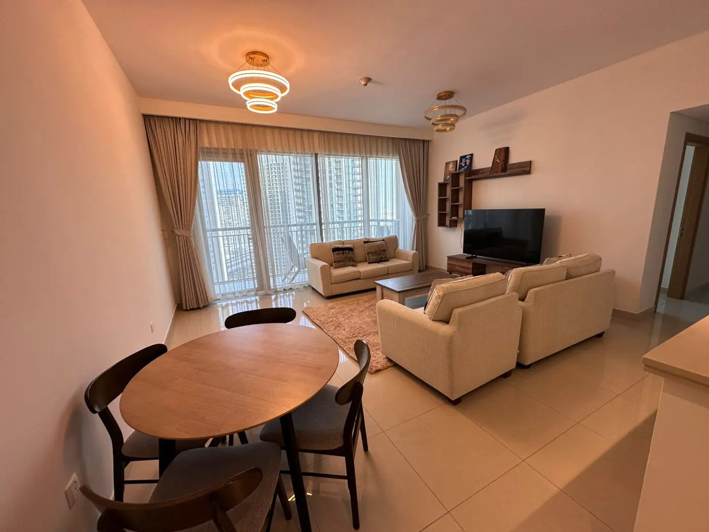 Bright apartment living room with a round wooden table, four chairs, and a cream-colored sofa set facing a large window with city views.