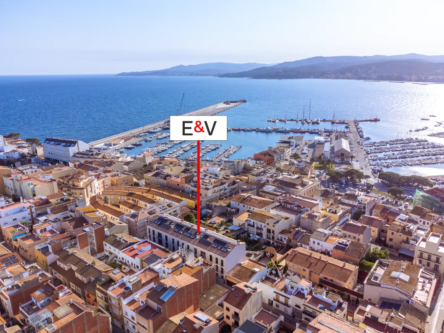 Aerial view of a coastal town with red-tiled roofs, a marina filled with boats, and a blue sea in the background. "E&V" marker.