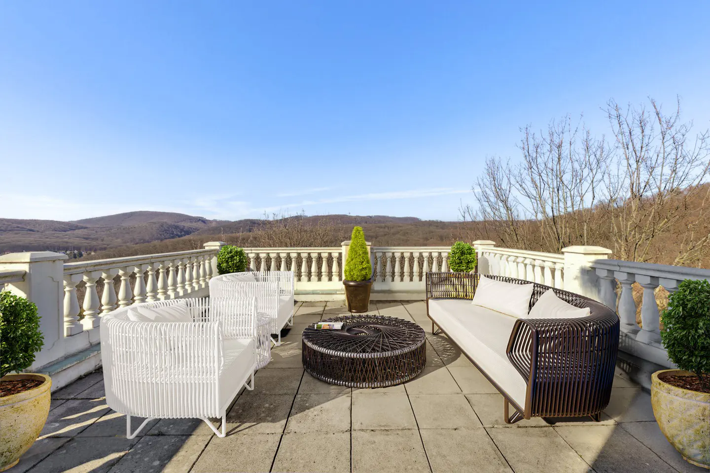 Outdoor patio with white wicker furniture, a brown wicker table, and potted plants. A white balustrade overlooks a mountain range under a blue sky.