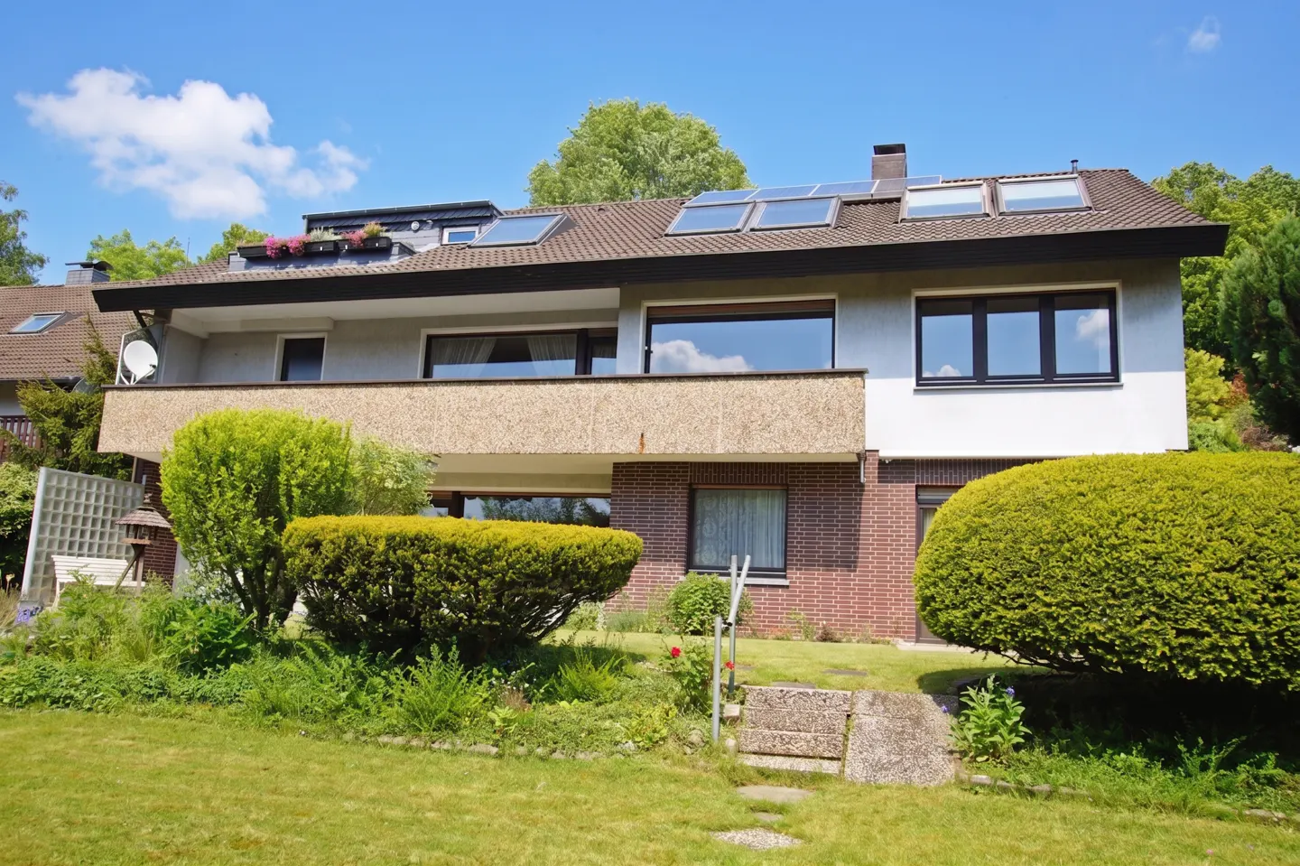 Two-story house with a balcony, brown roof, and solar panels. Green lawn and bushes in the front yard under a blue sky.