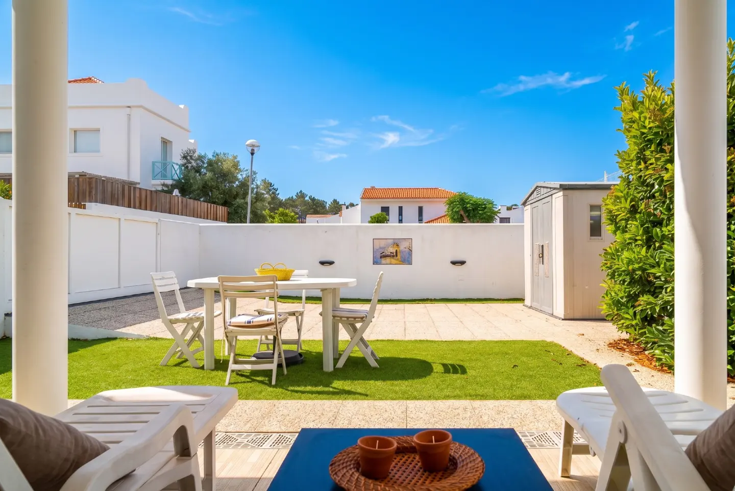 Outdoor patio with white table, chairs, and lounge chairs. Green grass, white walls, and blue sky in the background.