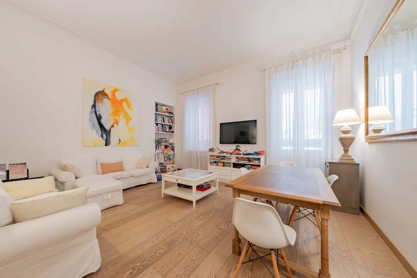Bright living room with hardwood floors, white sofas, a wooden dining table, and a large abstract painting on the wall.
