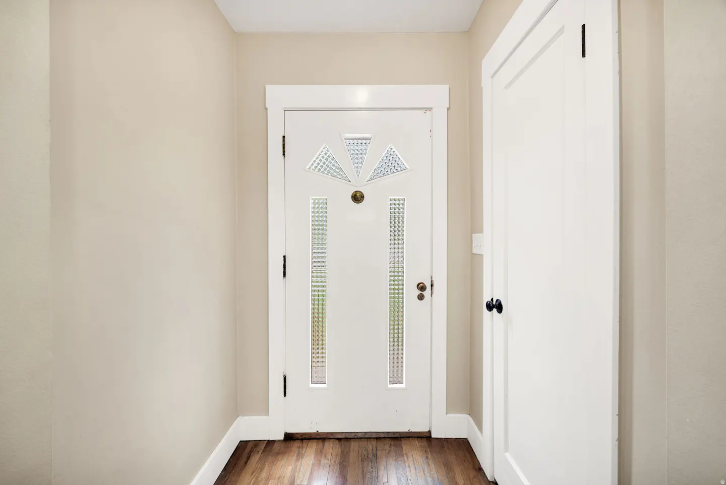 Entryway with a white door featuring decorative glass panels, a brass knob, and a white closet door to the right. Walls are beige, and the floor is wood.
