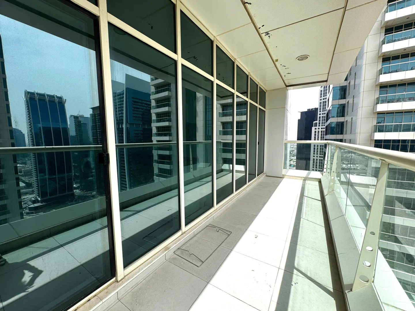 A bright balcony with white tile flooring, glass railings, and large windows reflecting city buildings.