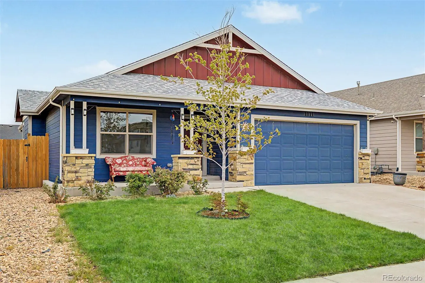Blue house with red trim, stone accents, and a blue garage door. A small tree sits in the green front lawn.