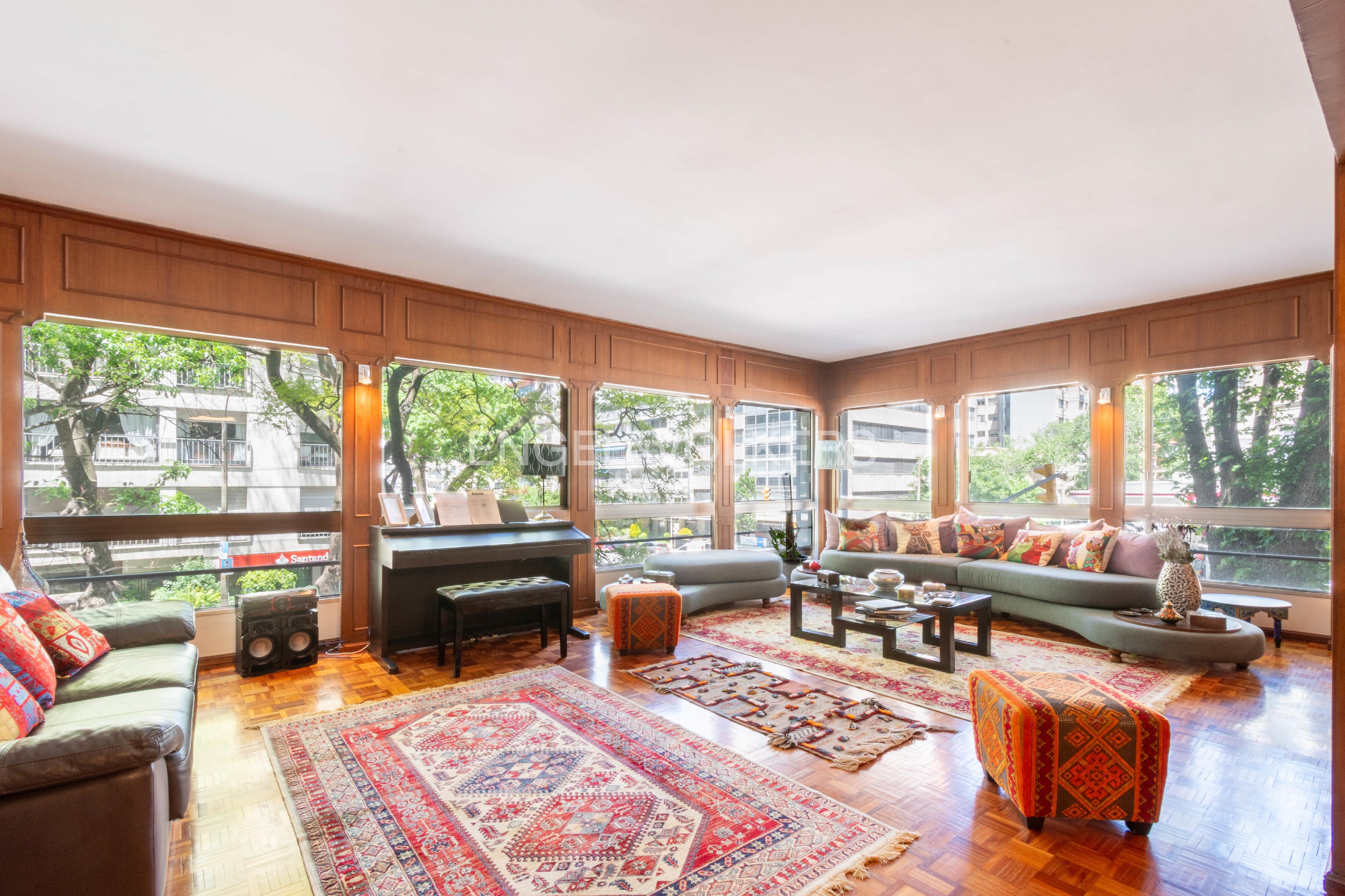 Spacious living room with wood-paneled walls, large windows, a piano, patterned rugs, and a curved sectional sofa.