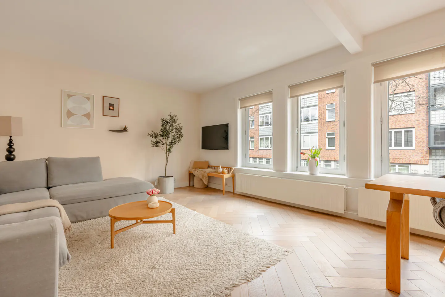 Bright living room with a gray sectional sofa, wood coffee table, and herringbone floors. Large windows offer a view of a brick building.