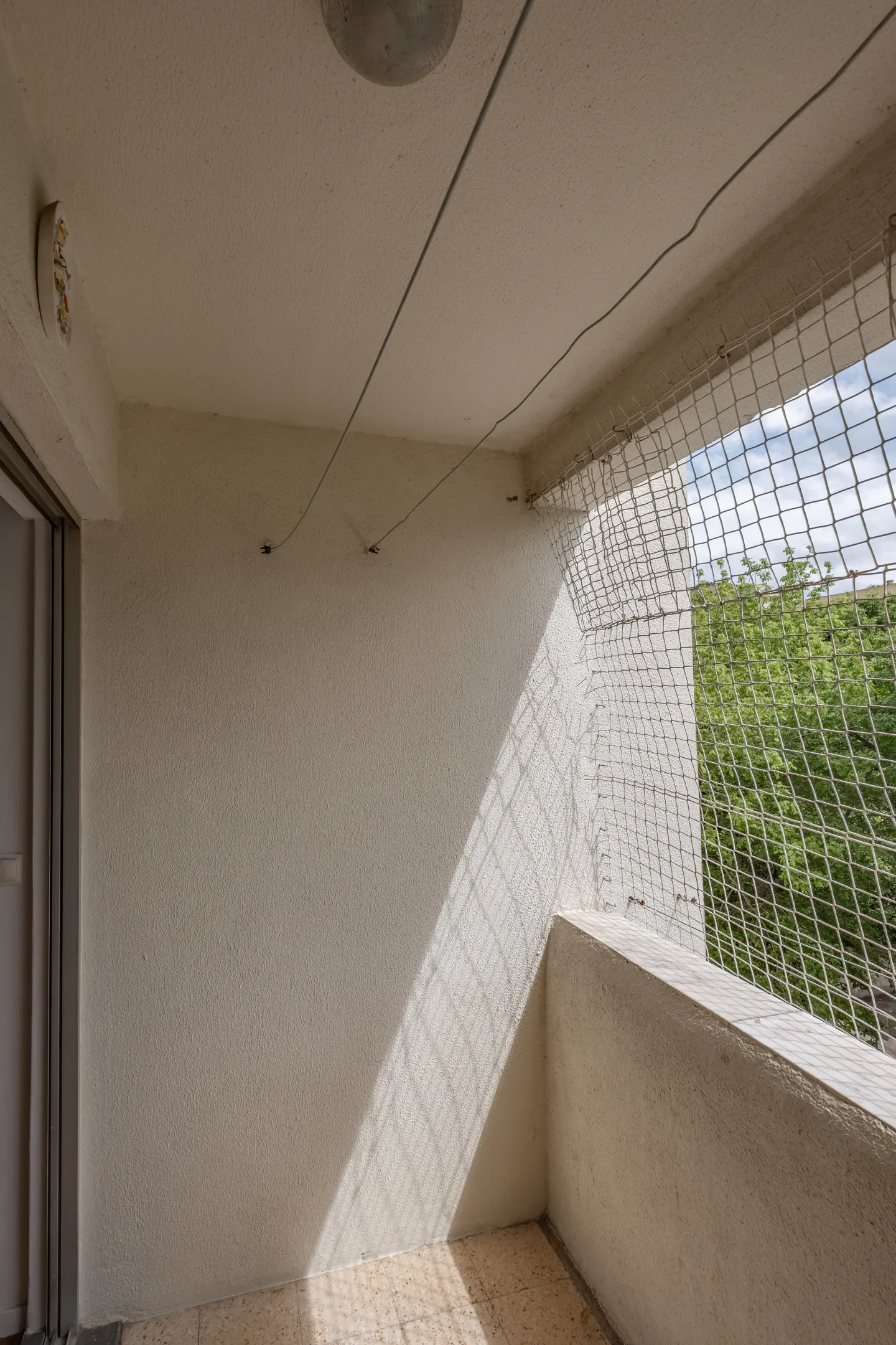 A bright, empty balcony with a white wall, a safety net, and a view of green trees.