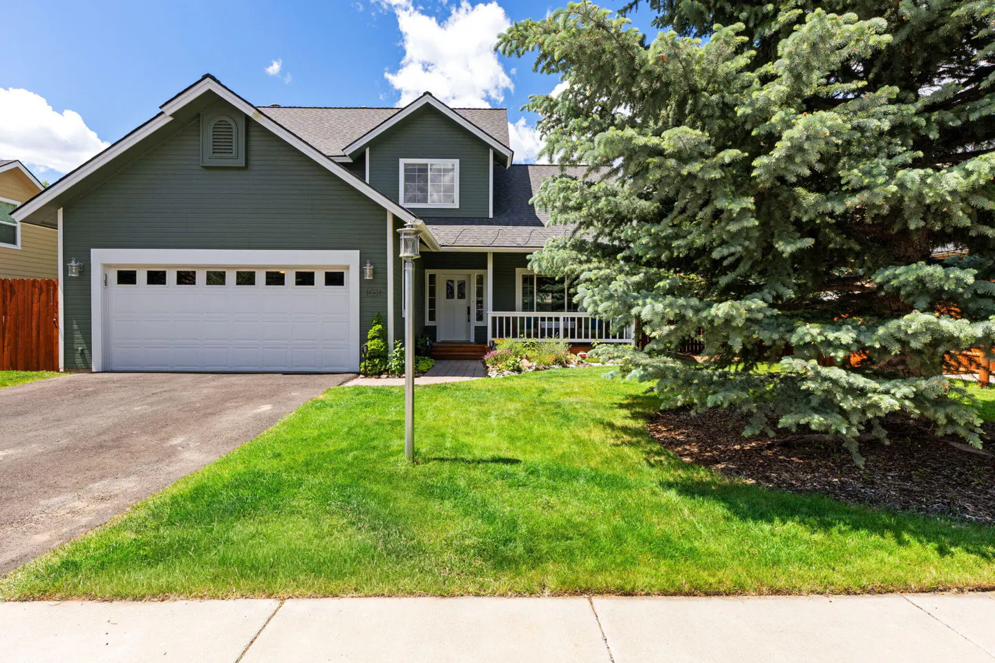 Two-story olive green house with a white garage door, front porch, and a large evergreen tree in the front yard.