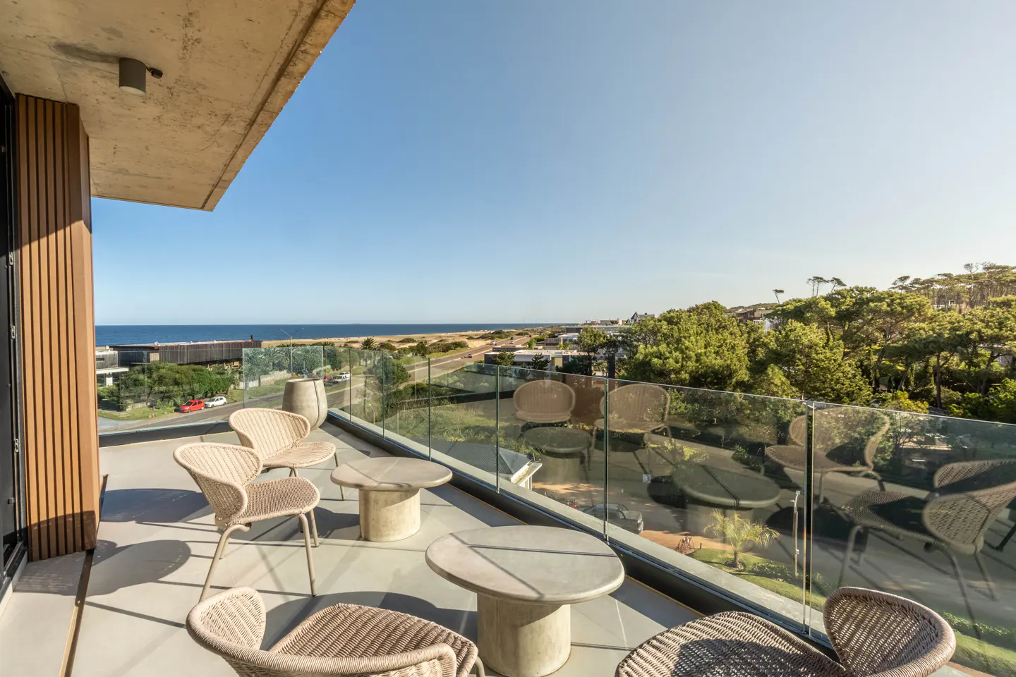 Balcony with wicker chairs and tables overlooks ocean and trees under a blue sky. Glass railing surrounds the outdoor space.